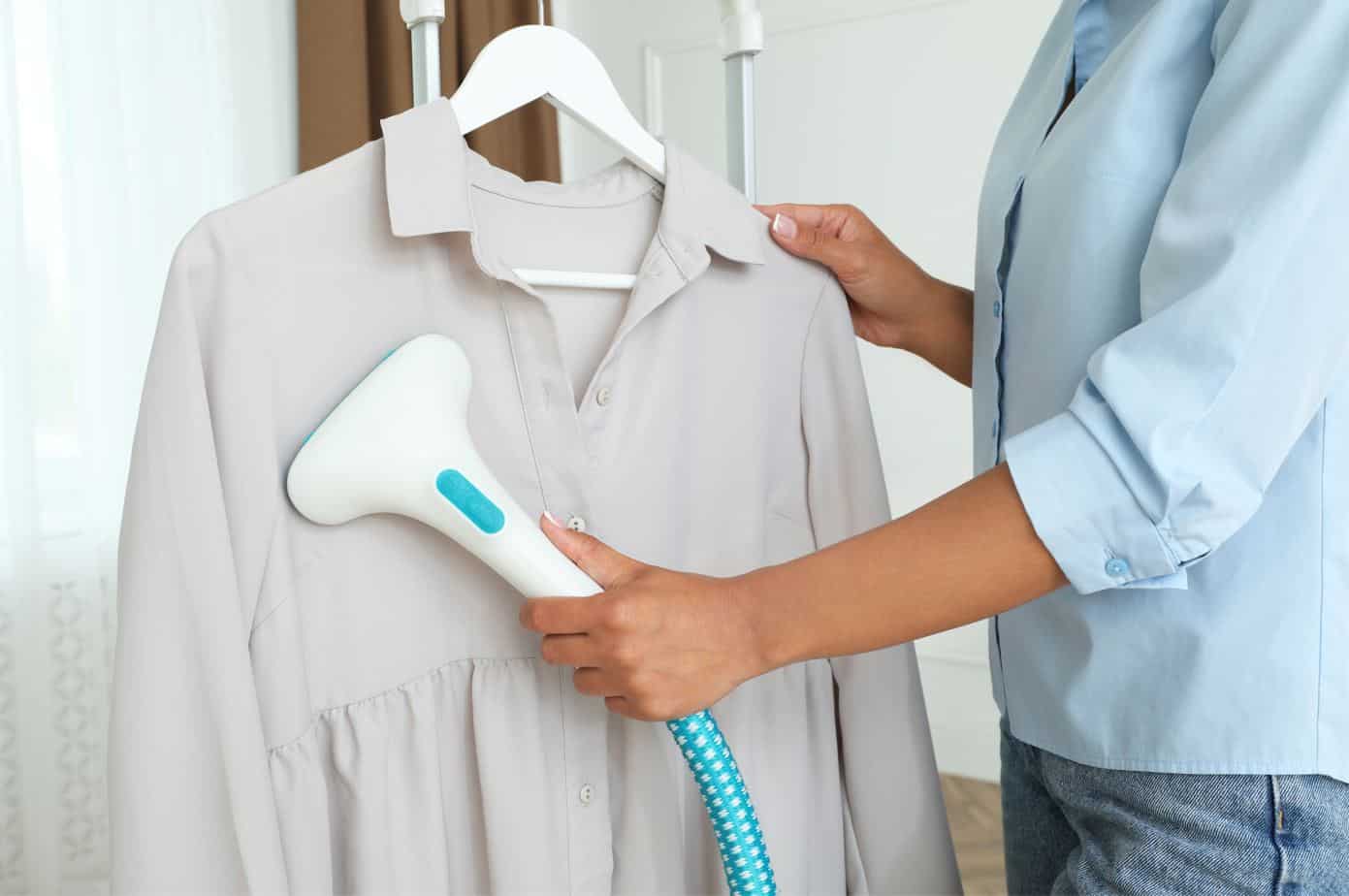woman wearing blue button up shirt using a white steam iron to iron a hanging grey blouse