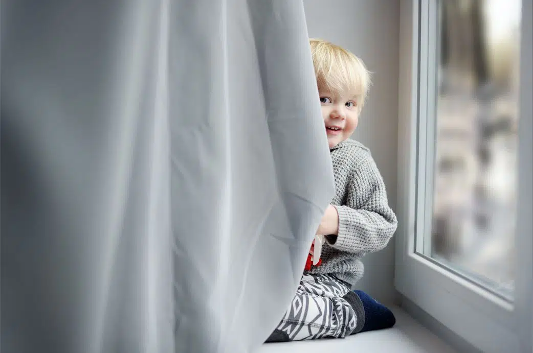blonde toddler hiding behind grey fabric curtains