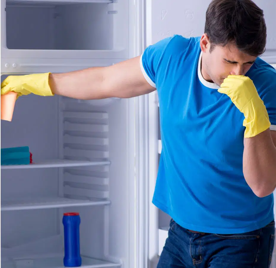 man wearing blue shirt holding his nose with open empty fridge in the background
