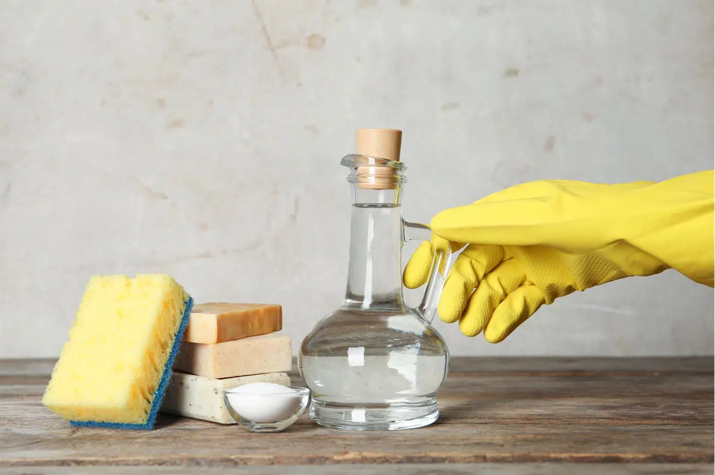 person wearing yellow gloves holding a bottle of vinegar next to a sponge and assorted hand soaps