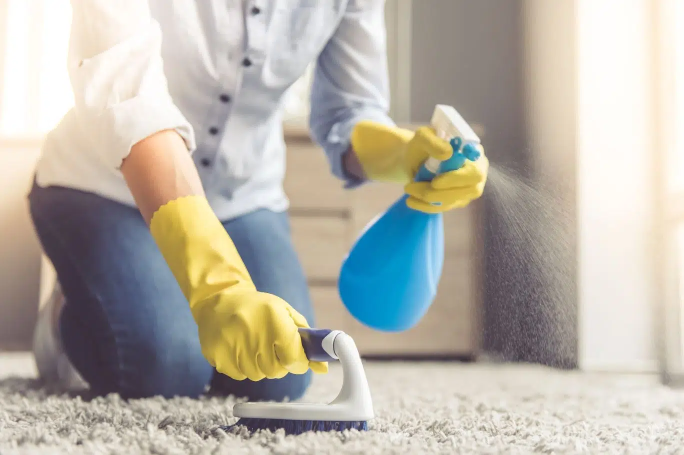 woman wearing yellow rubber gloves spritzing cleaning chemical onto her gray carpet